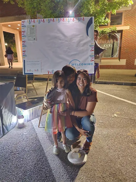 Woman and two children smiling in front of a welcoming event board at night.
