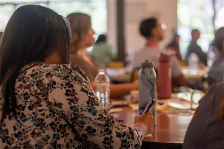 People attending a workshop, taking notes around a table with water bottles, in a bright room.