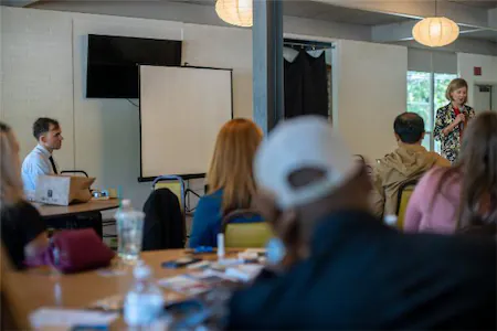 Speaker presenting to an audience in a conference room with a projector and screen.