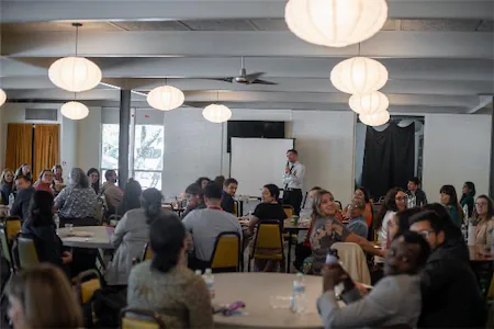 People listening to a speaker at a conference in a brightly lit room with round tables.