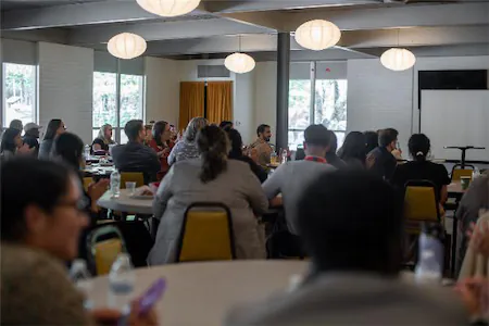 Audience attentively listens during a conference in a well-lit room with round tables and hanging lights.