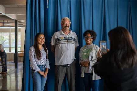 Three people smiling for a photo indoors with a blue curtain backdrop, while another person takes a picture with a phone.