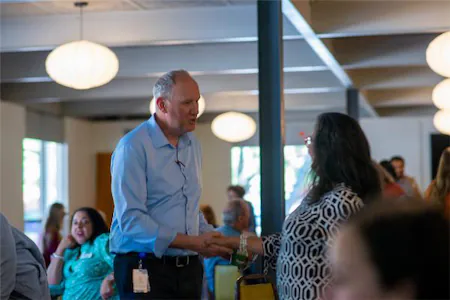 Man and woman greeting each other at a social event in a well-lit room, surrounded by people and round lights.