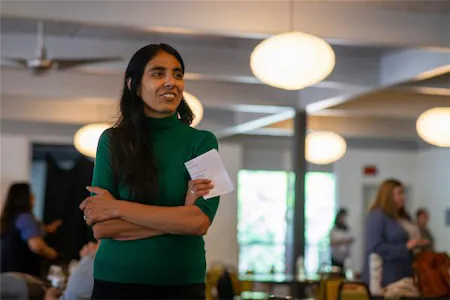 Woman in green sweater holding paper, standing in a brightly lit room with people in the background.