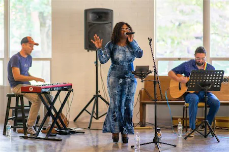 Musicians performing live: singer with mic, keyboardist, and guitarist in a bright indoor setting.