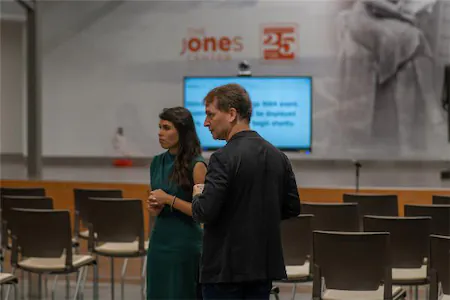 Two people conversing in a conference room with empty chairs and a screen in the background.
