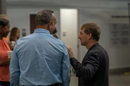 Two men engaged in conversation at an indoor event, with people in the background.