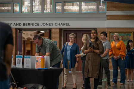 People standing in line at an event in front of a chapel entrance, engaging with various informational booths.