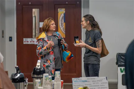 Two women chatting at a coffee station during a social event, one holding a drink and the other a snack.