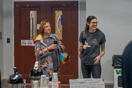 Two women chatting at an event, one holding a drink, the other enjoying a snack, with a table of refreshments nearby.