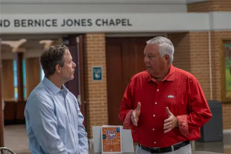 Two men having a conversation inside a chapel hallway, one wearing a red shirt and the other a light blue shirt.