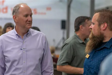 Two men engaged in conversation at an indoor event, surrounded by people in casual attire.