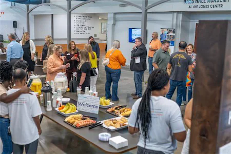 Community gathering at indoor venue with people socializing around a table of refreshments and snacks.