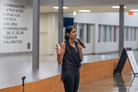 A person speaking into a microphone at an indoor venue, holding a phone, with directional signs in the background.