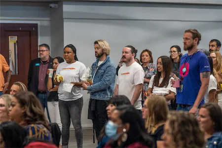 A diverse group of people attentively listening at a community event, some holding drinks and snacks.