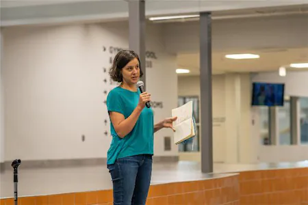 Person speaking on a stage, holding a microphone and papers, in a well-lit indoor setting.
