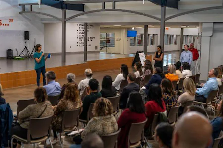 Audience listening to a speaker at an indoor seminar with a stage and presentation setup.