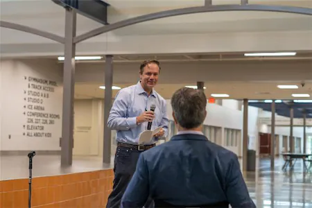 Man speaking at an indoor event with microphone, facing audience in spacious venue.