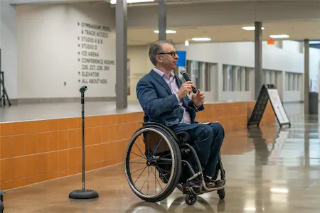 Person in wheelchair speaking into microphone indoors, near a directional sign and another mic stand.