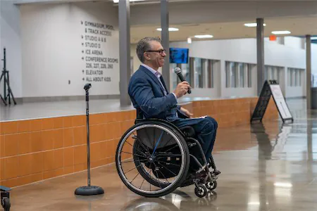 Person in a wheelchair giving a speech indoors, holding a microphone, with a hallway and directional signs visible.