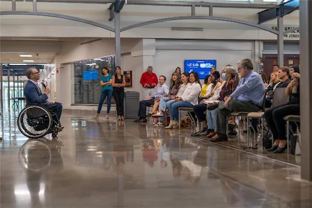 A speaker in a wheelchair addresses a seated audience in a modern hall, engaging in a discussion or presentation.