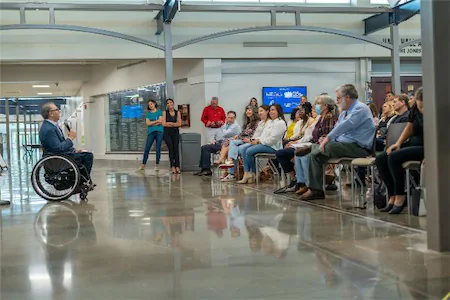 Speaker in a wheelchair addressing a seated audience in a modern hall setting.