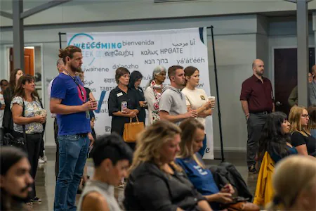 Diverse group of people attending a welcoming event in a large hall, with multilingual welcome sign in the background.