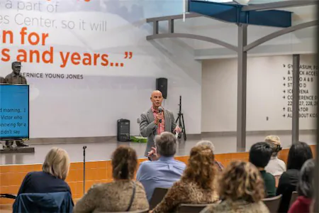 Speaker addressing an audience in a modern conference room with a large quote displayed on the wall behind.