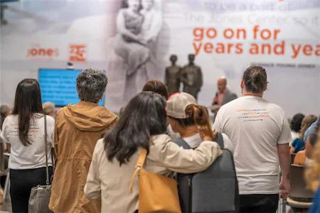 Audience attending a presentation in a large room, with a speaker and a screen visible in the background.