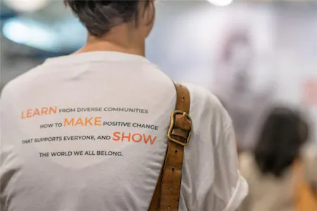 Person wearing a white shirt with motivational text about diversity and positive change, blurred background, brown strap.