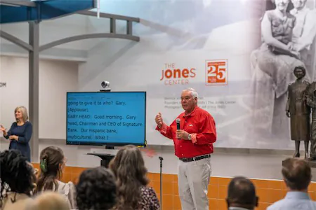 Speaker addressing audience at The Jones Center event with presentation screen and statue in background.