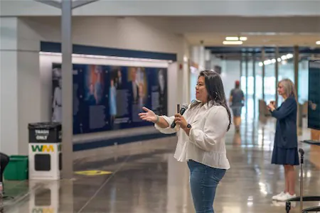 Woman speaking with a microphone in a hallway setting, with a person in the background taking a photo.