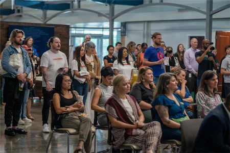 Audience attentively listens during an indoor event, diverse group sitting and standing in a spacious hall.
