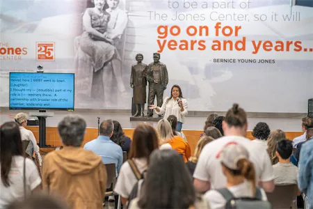 Speaker presenting at the Jones Center with an audience, backdrop features quote and statue.