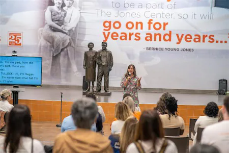Person speaking at an event in front of an audience with a statue and quote in the background.