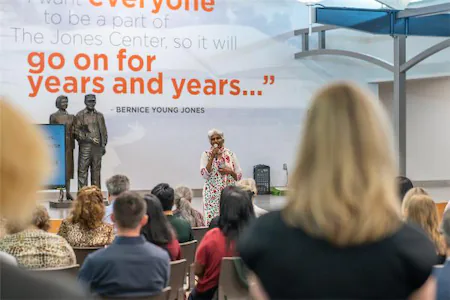 Speaker addressing audience at indoor event, with inspirational quote on wall in background.