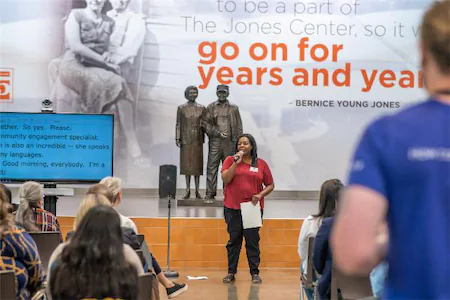 Woman speaking at a community center event, people seated, large poster in the background.