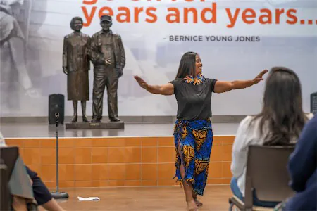 A woman dances during a performance, with seated audience and historical statues in the background.
