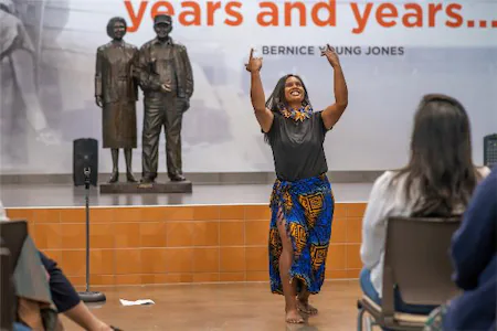 Performer in vibrant attire dances joyfully at an indoor event, with an audience watching and statues in the background.