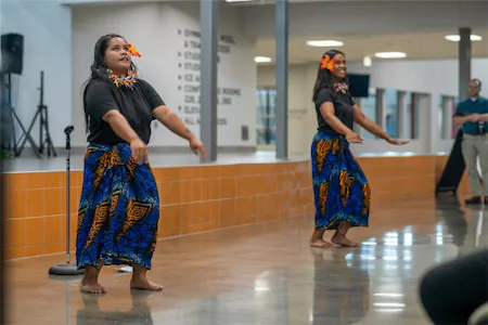 Two performers in traditional Pacific island attire dancing in a community center.