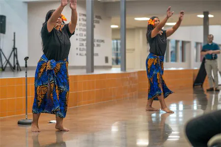 Two dancers in traditional attire perform a cultural dance indoors, with vibrant blue skirts and orange floral accents.