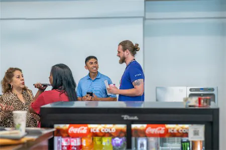 People socializing near a beverage cooler, holding cups and engaging in conversation at a casual gathering.