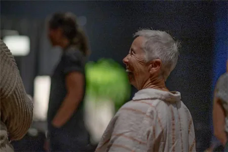A smiling elderly woman with short hair enjoys a social gathering, highlighted against a blurred background.
