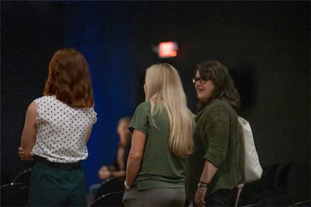 Three women standing and chatting in a dimly lit room during an event.