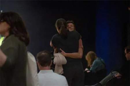 Two people happily embracing in a busy indoor setting with others in the background.