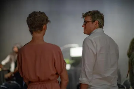 Two people talking in a dimly lit room, one wearing a white shirt, the other in a pink dress.