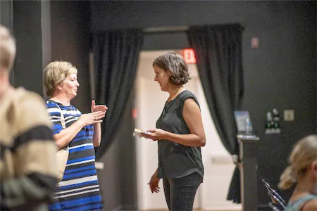 Two women conversing at an indoor event, one holding a plate, with a curtain backdrop.