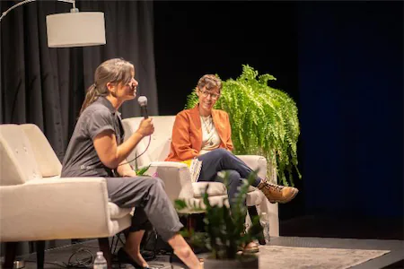 Two people seated on a stage engaging in a conversation, with a microphone and a green fern plant in the background.