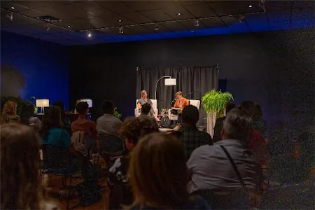 Audience attending an intimate live talk show in a dimly lit studio with two speakers on stage.