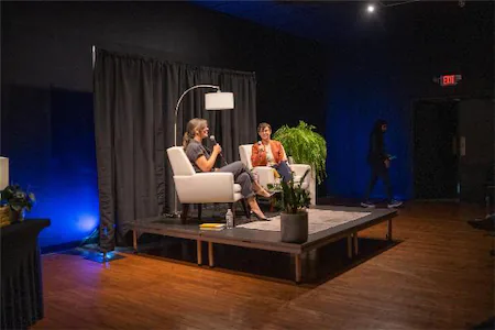 Two women engaged in a panel discussion on a stage with plants and a dark curtain backdrop.